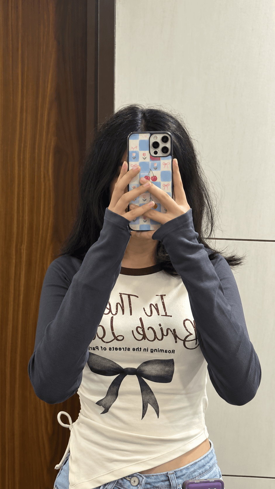 Mirror selfie featuring a pastel blue checkerboard phone case with cherries, bows and tiny flowers.